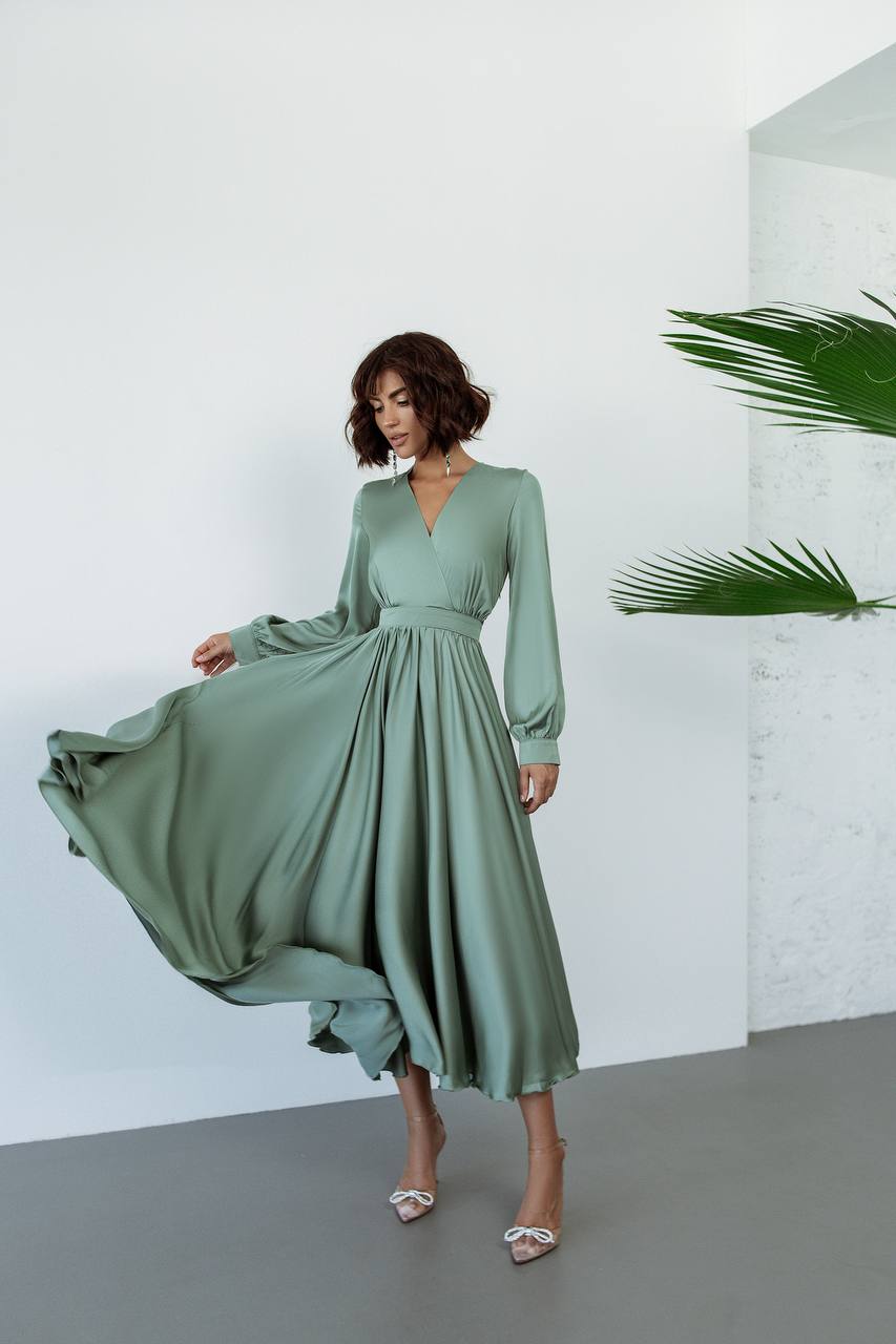 Woman wearing a green dress standing against a white wall with a plant.