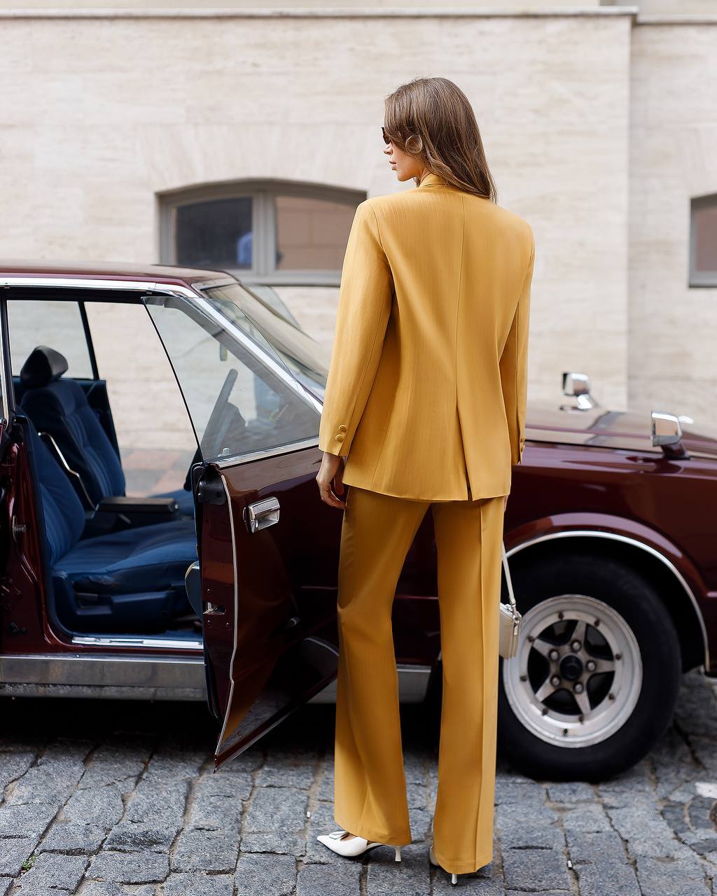 Woman in a mustard yellow suit standing next to an open car door on a street.