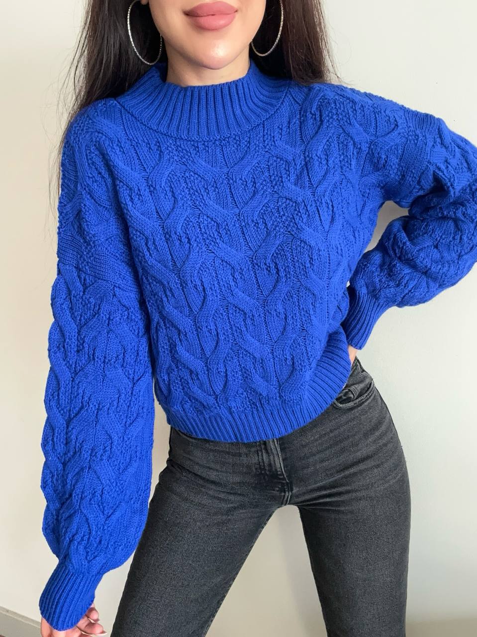 Person wearing a blue cable knit sweater and dark jeans against a plain background
