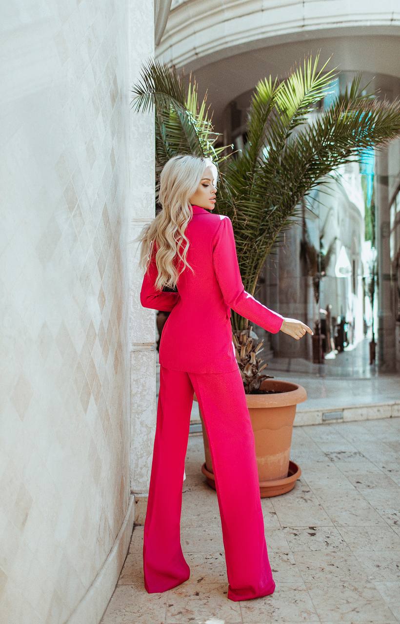 Woman in a bright pink suit standing against a white wall with plants in the background