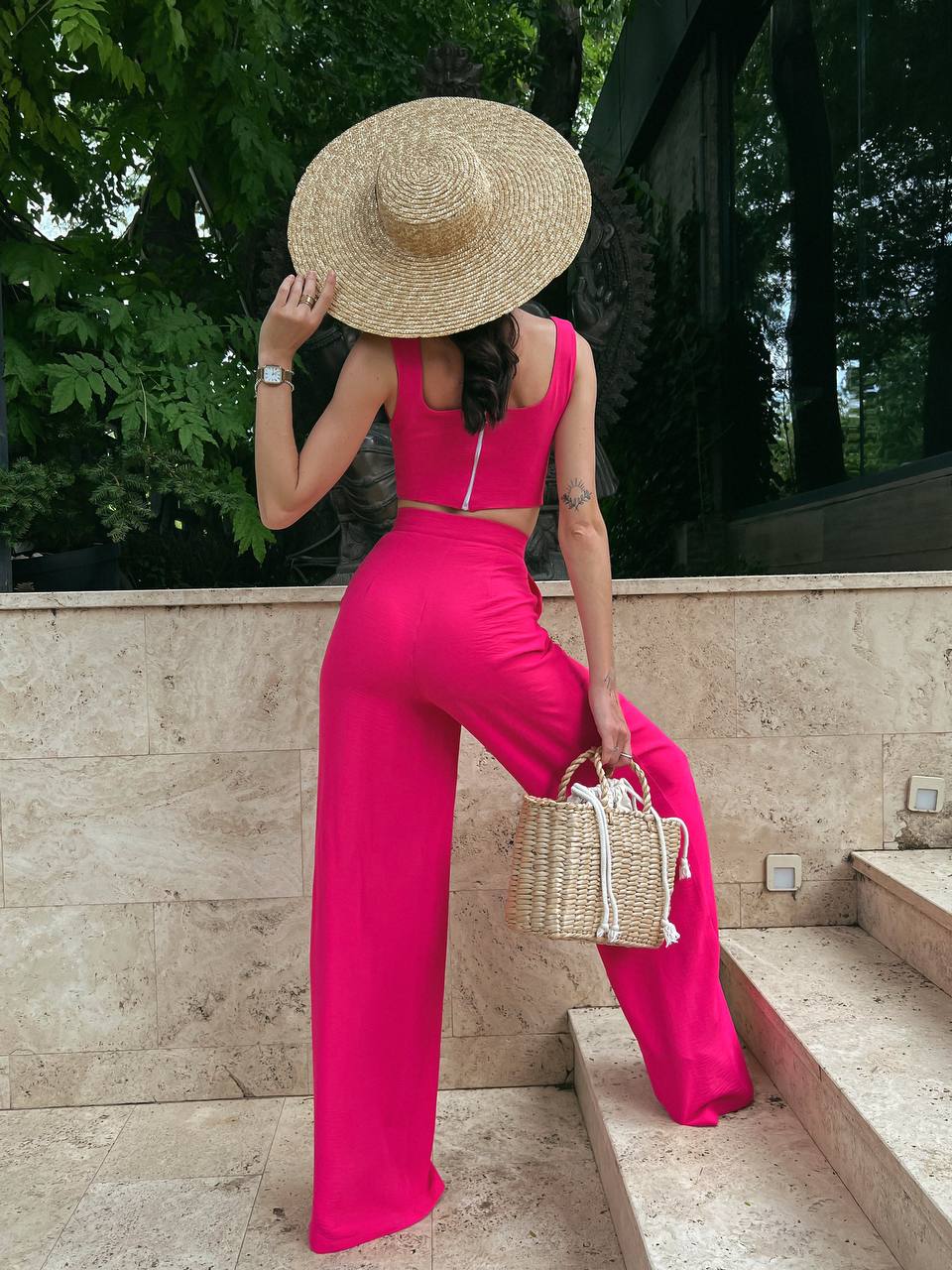 Woman in a bright pink jumpsuit and straw hat holding a small bag, standing outdoors.