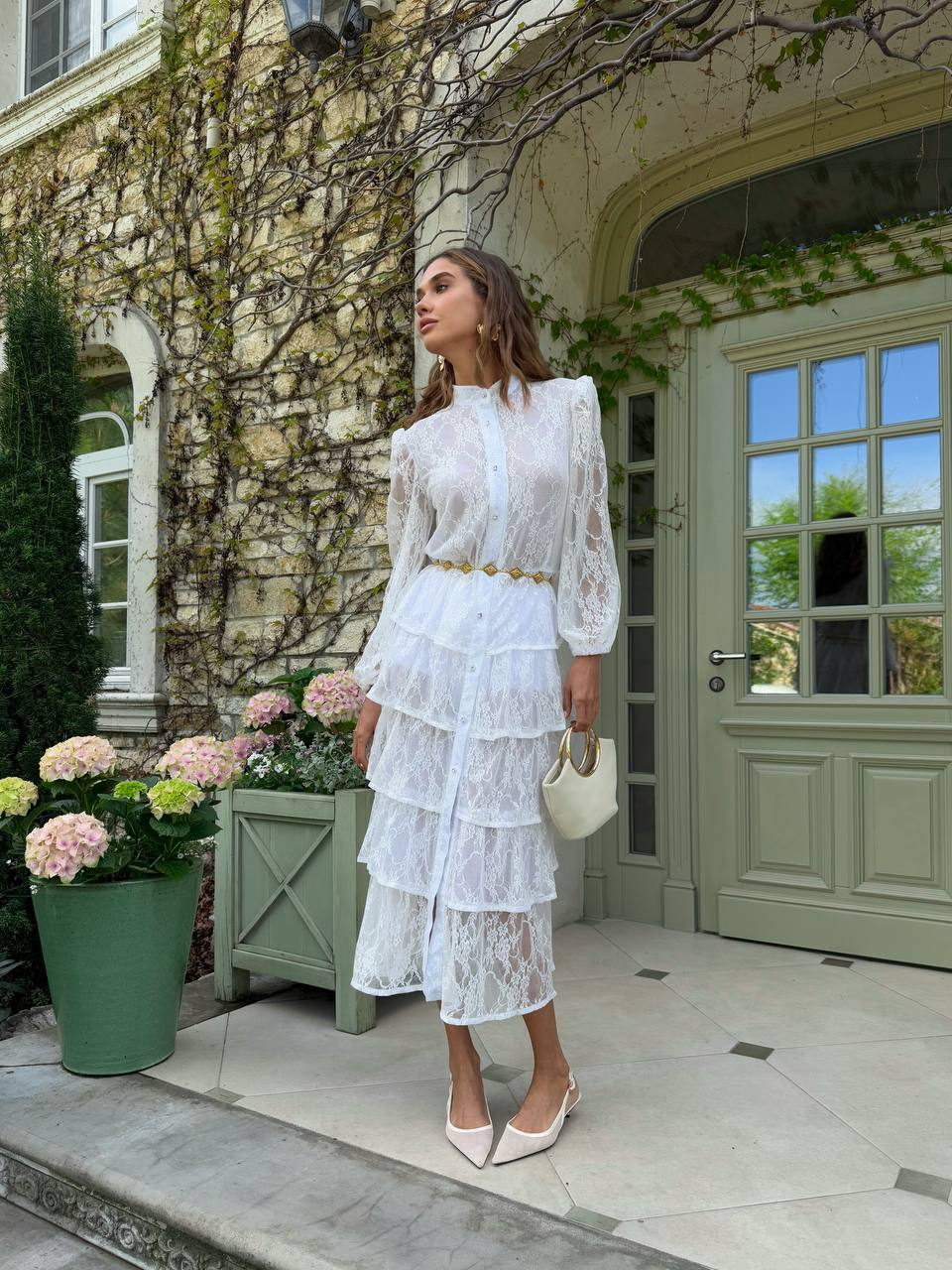Woman in a white lace outfit standing in front of a stone building with greenery.