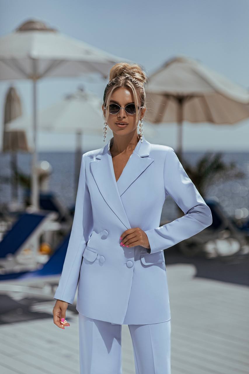 Woman in a white suit standing on a beach with umbrellas and lounge chairs in the background.