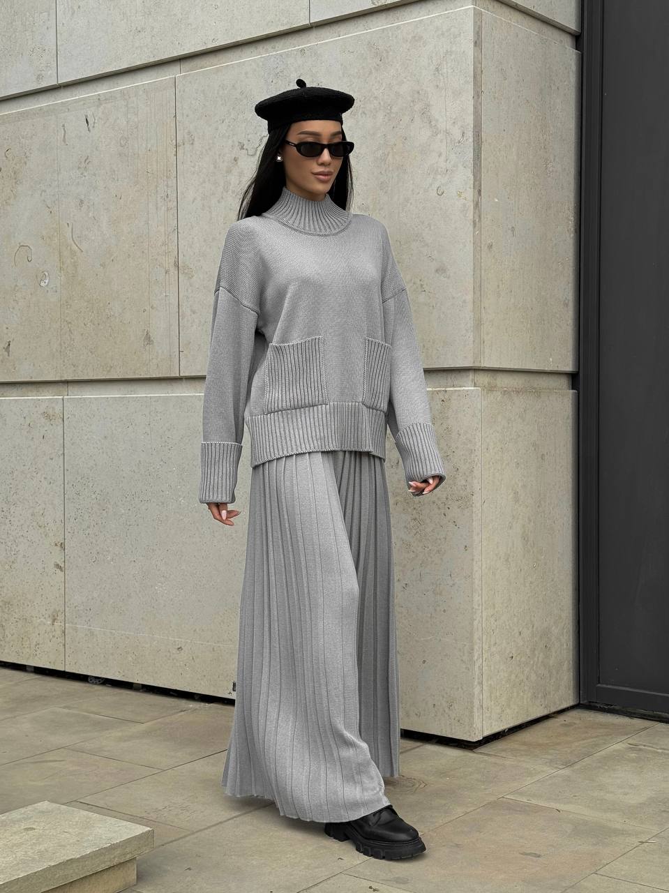 Person wearing a gray outfit with a black beret and sunglasses, standing against a stone wall.