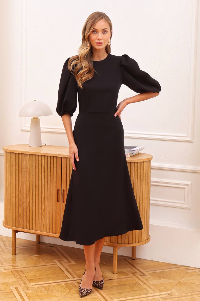 Woman wearing a black dress standing in a room with wooden furniture.