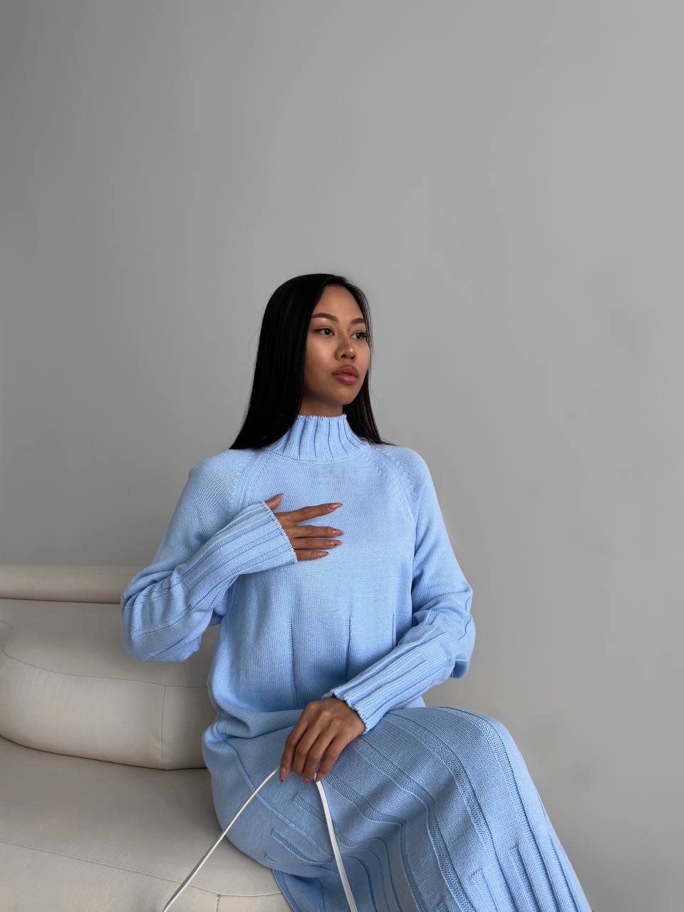 Woman wearing a light blue sweater and skirt sitting on a white couch against a gray wall.
