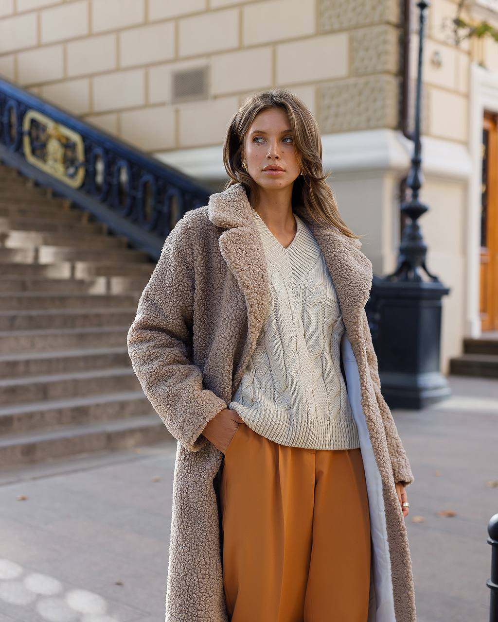 Woman wearing a long beige coat, light sweater, and mustard pants on a city street.