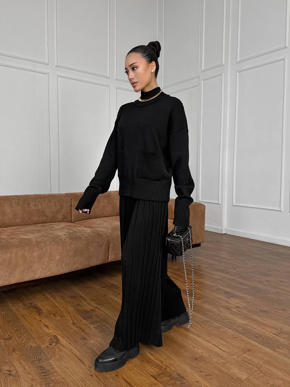 Woman in a black outfit standing in a room with a brown couch and white paneled walls.