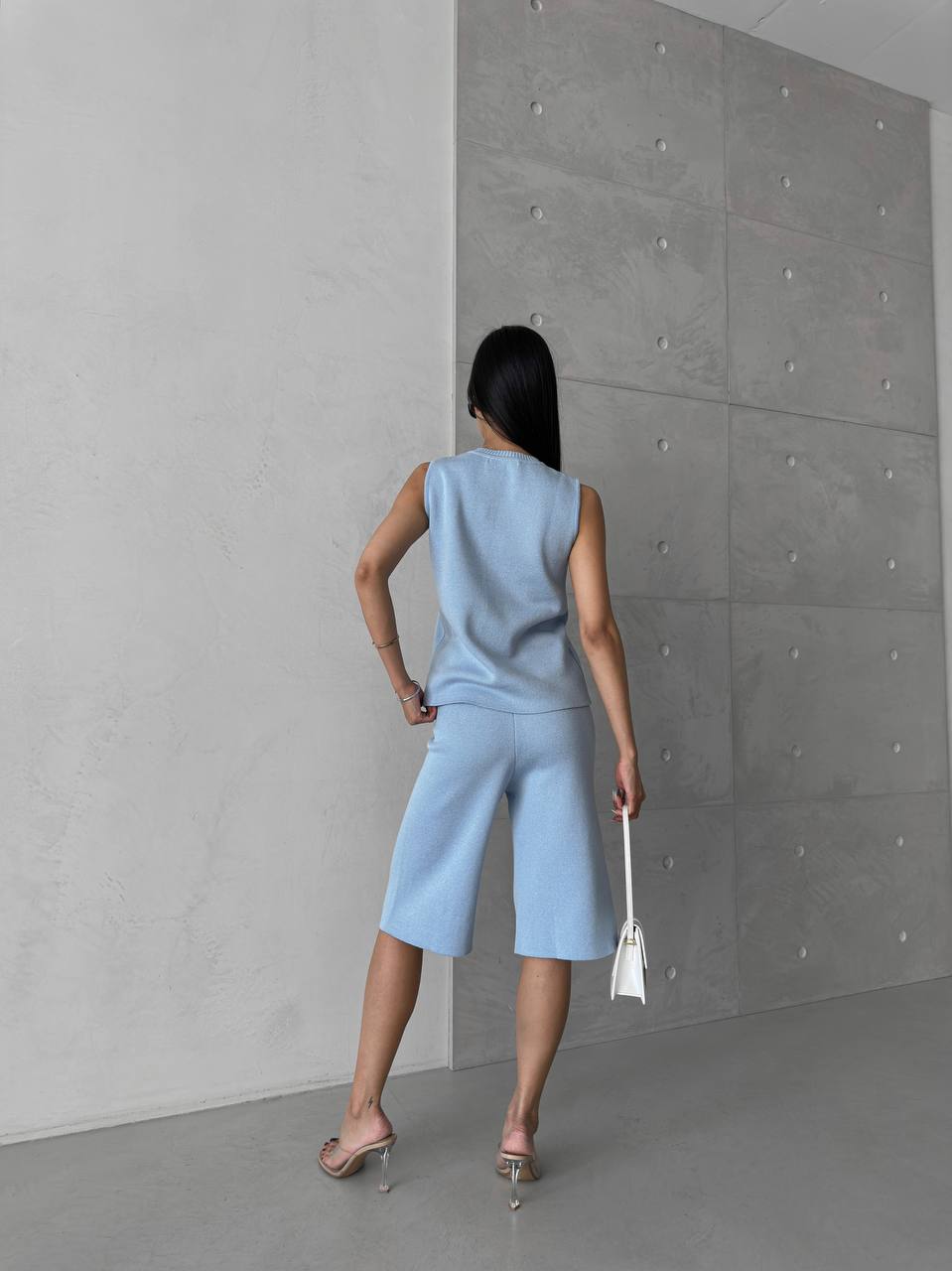 Woman in a light blue sleeveless top and shorts standing against a gray concrete wall.