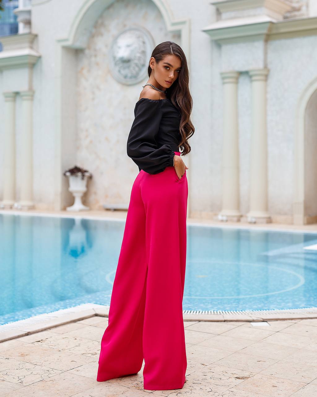 Woman in black top and pink pants standing by a pool with classical architecture in the background