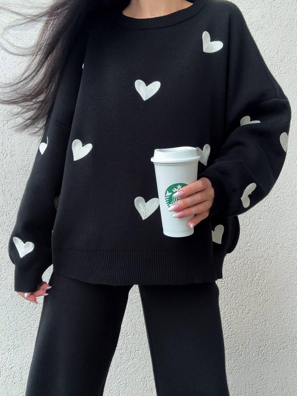 Person wearing a black sweater with white heart patterns, holding a Starbucks cup.