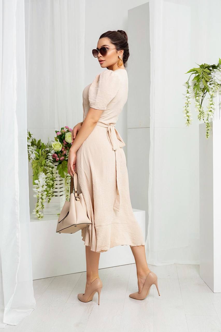 Woman in a beige dress standing in a minimalistic room with white walls and a plant.
