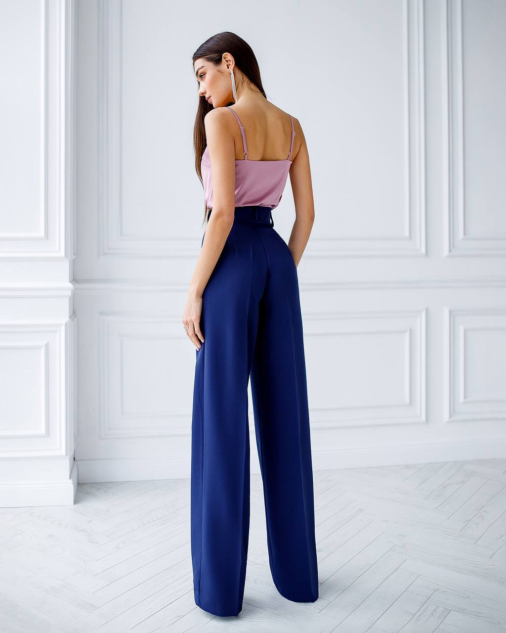 Woman wearing a pink top and blue high-waisted pants in a white room.