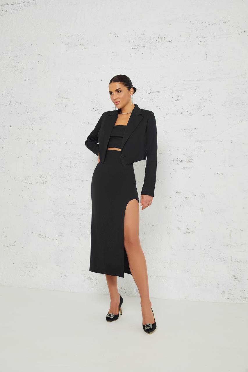 Woman wearing a black outfit with a high slit skirt against a white brick wall.