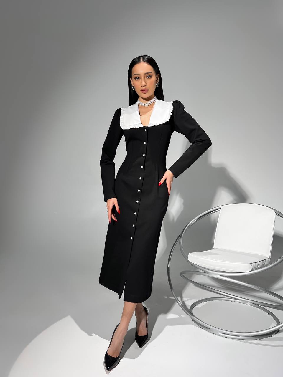 Woman in a black dress with white collar standing next to a modern chair on a gray background