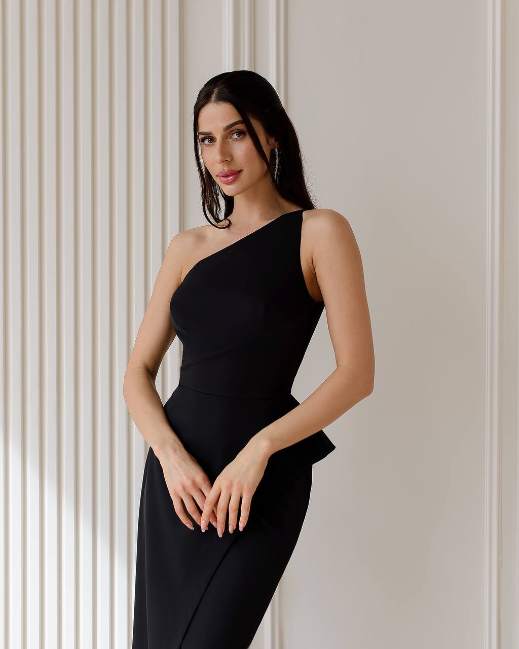 Woman wearing a black one-shoulder dress against a neutral wall.