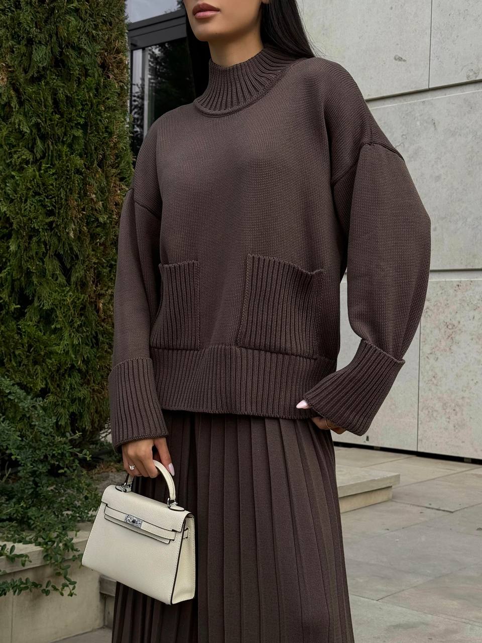 Person wearing a brown sweater and skirt holding a white handbag outdoors.