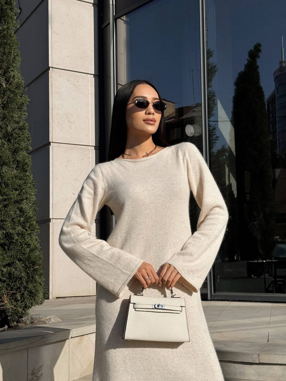 Woman in a light-colored dress holding a matching handbag, standing in front of a reflective building.