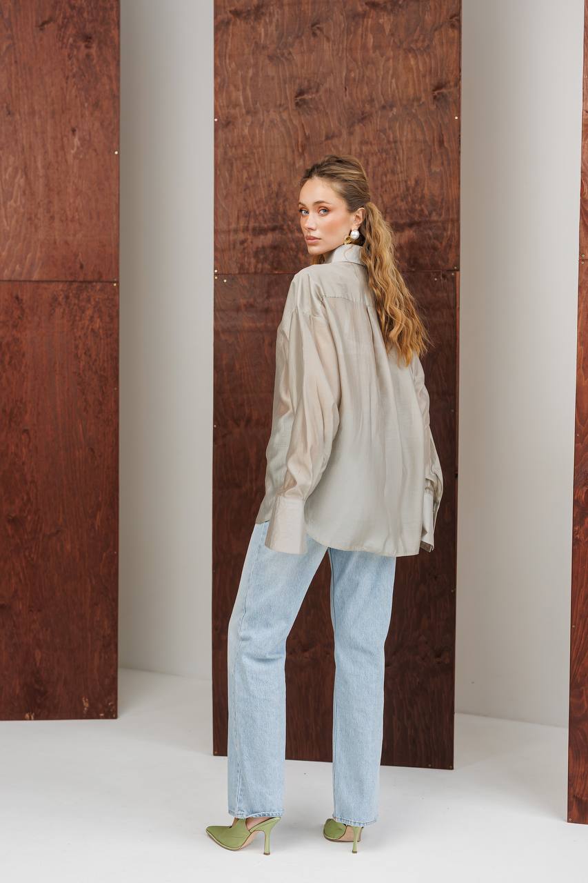 Woman wearing a light-colored shirt and blue jeans standing against a wooden panel.