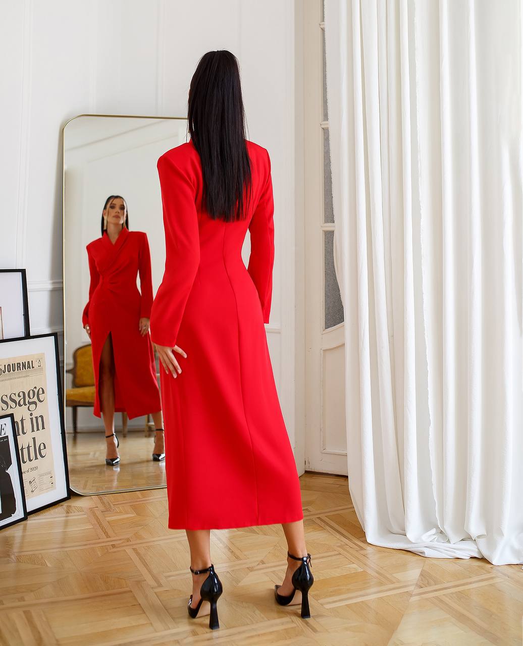 Woman in a red dress standing in front of a mirror in a room with white walls and curtains.