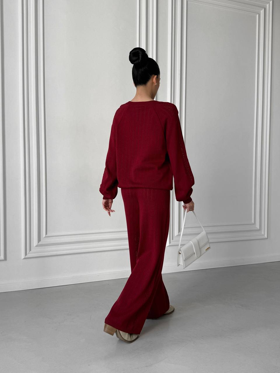 Person wearing a burgundy outfit holding a white handbag against a white paneled wall.