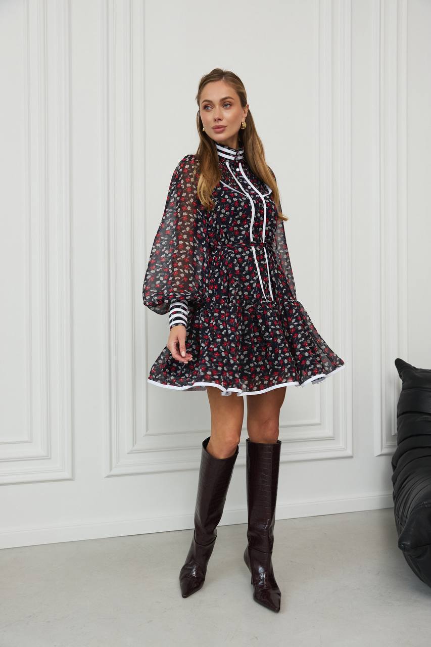 Woman wearing a floral dress with long sleeves and knee-high boots in a room with white paneled walls.