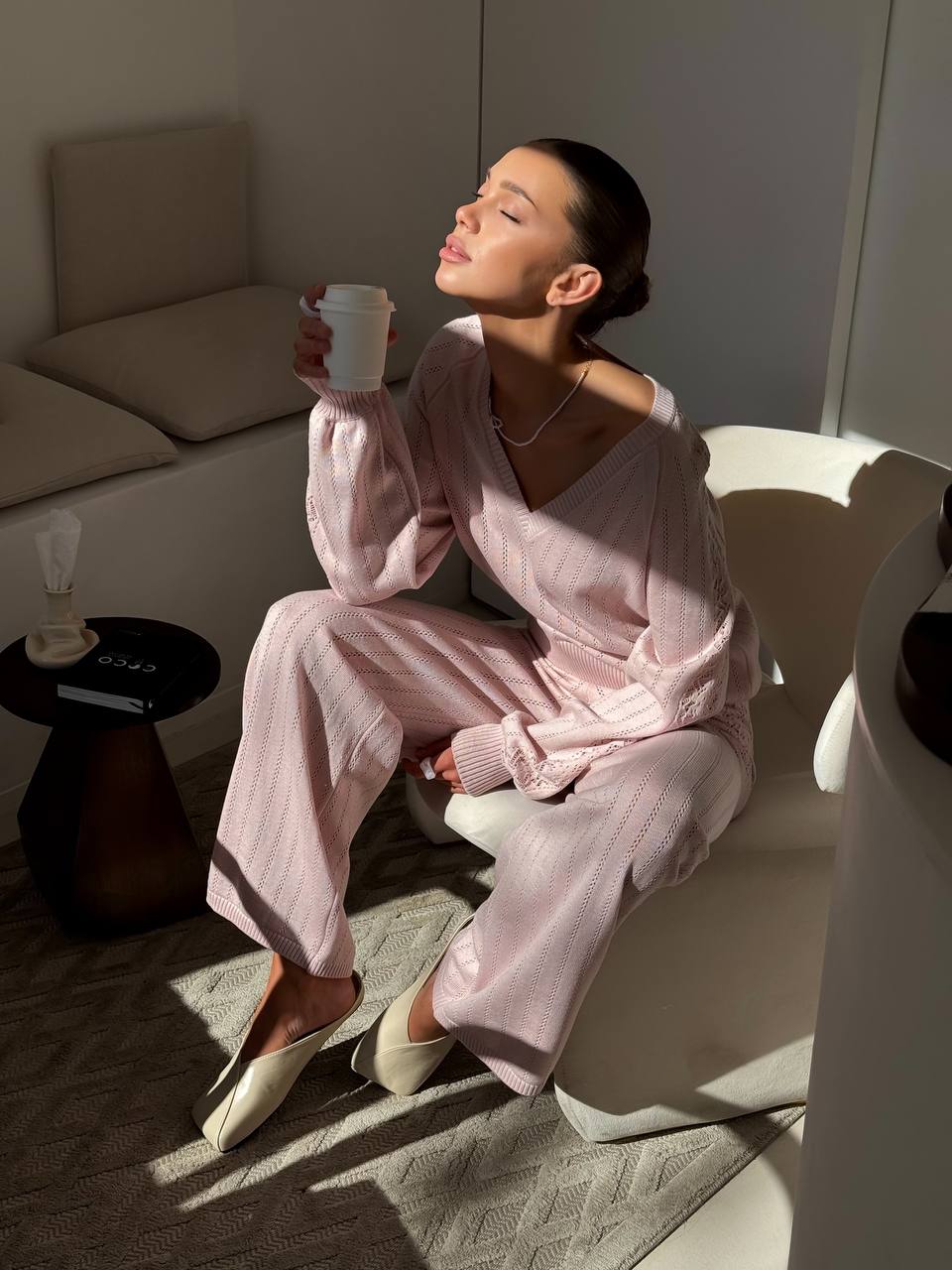 Woman in pink pajamas holding a cup in a cozy room with sunlight streaming in.