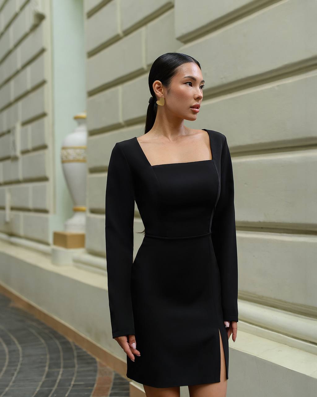 Woman wearing a black dress with long sleeves and a square neckline, standing against a light-colored wall.