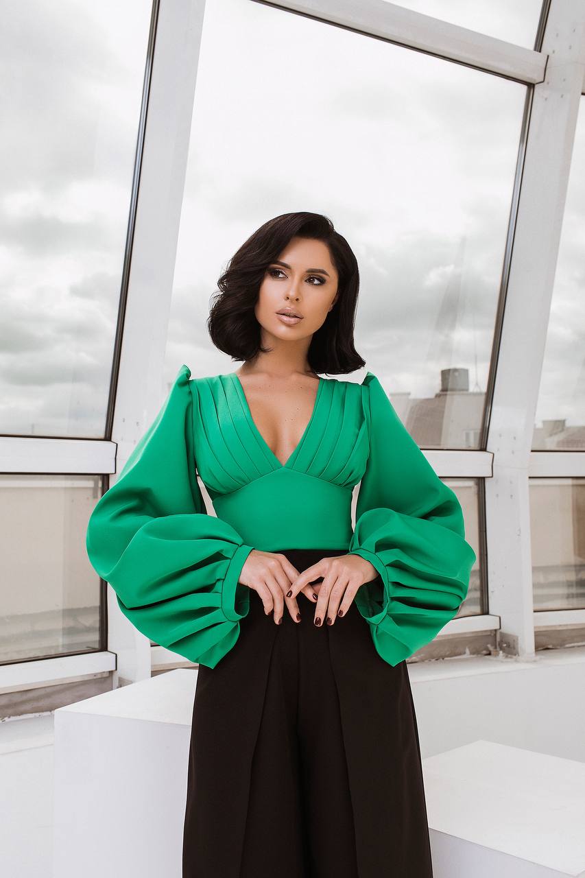 Woman wearing a green top with wide sleeves and black pants in an indoor setting.