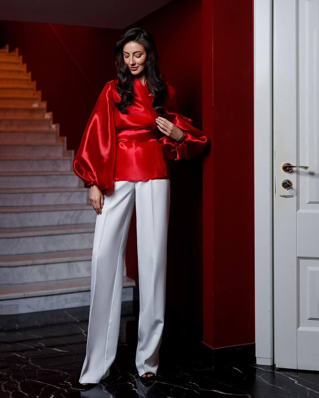 Woman in a red satin top and white pants standing in a room with a staircase.