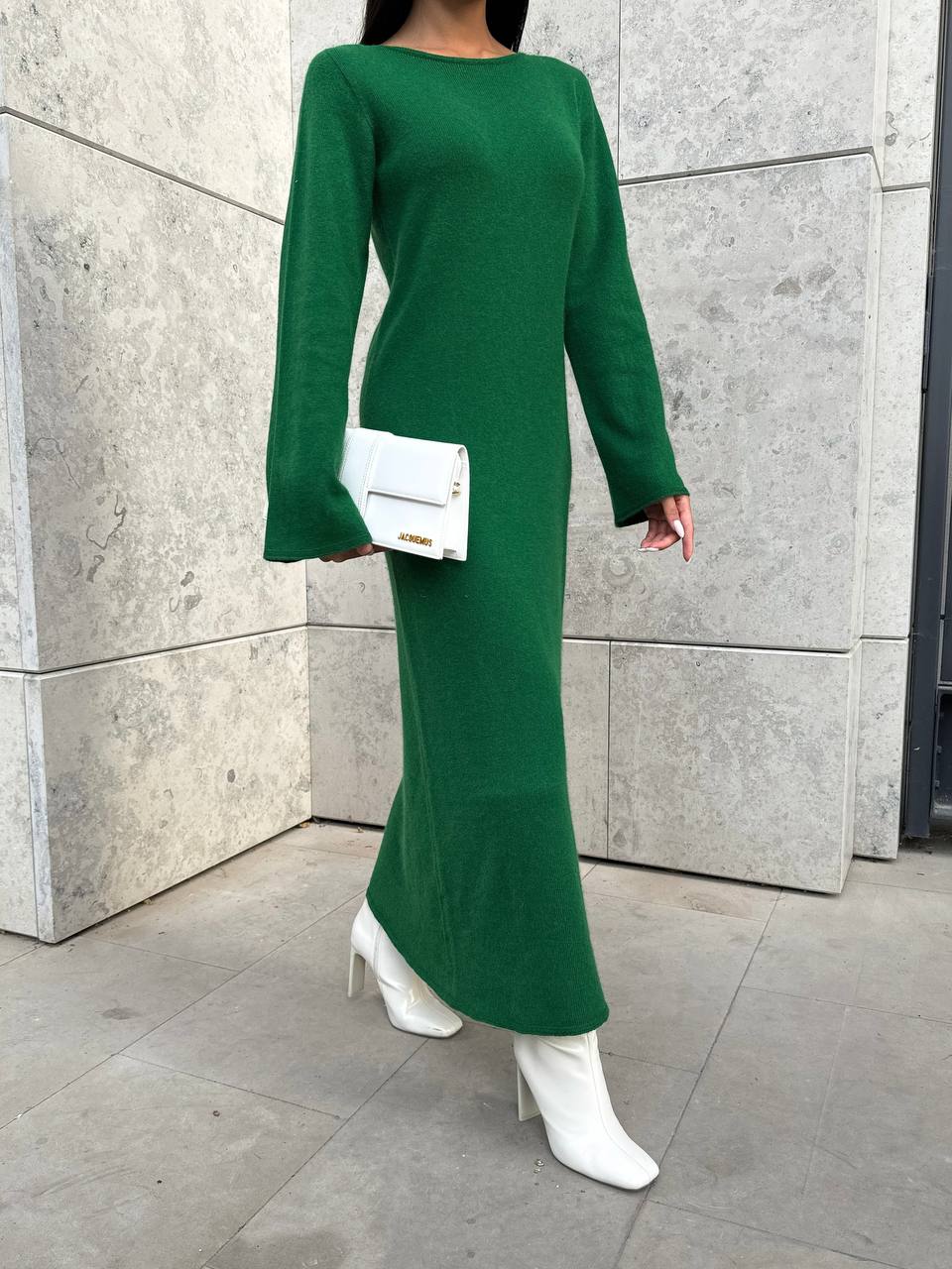 Person wearing a green dress with white boots and a white clutch against a gray wall.