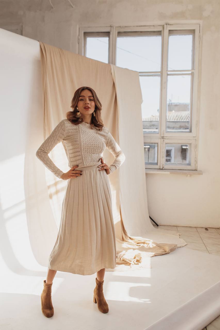 Woman in a beige dress standing in a room with large windows and draped fabric.