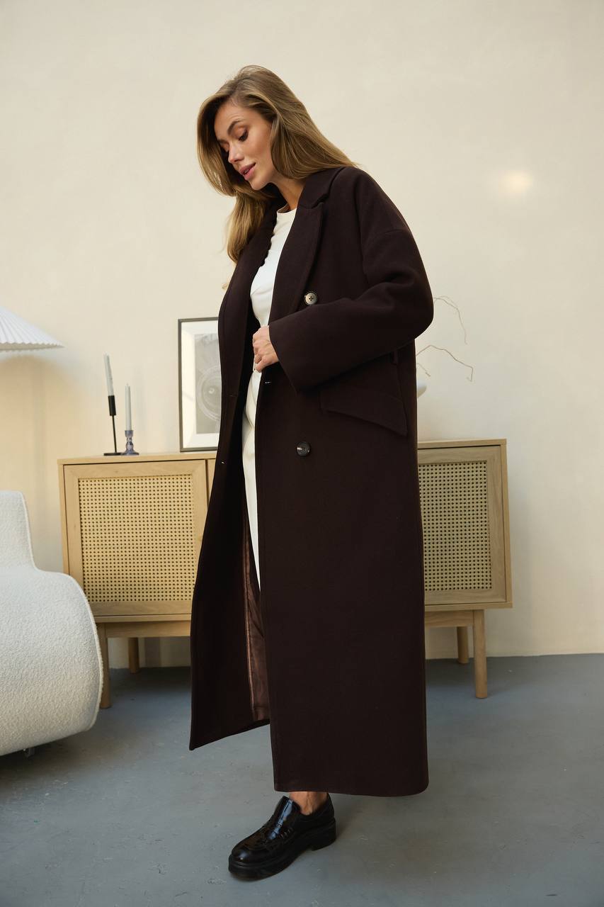 Woman wearing a long dark brown coat in a room with a light-colored wall and furniture.