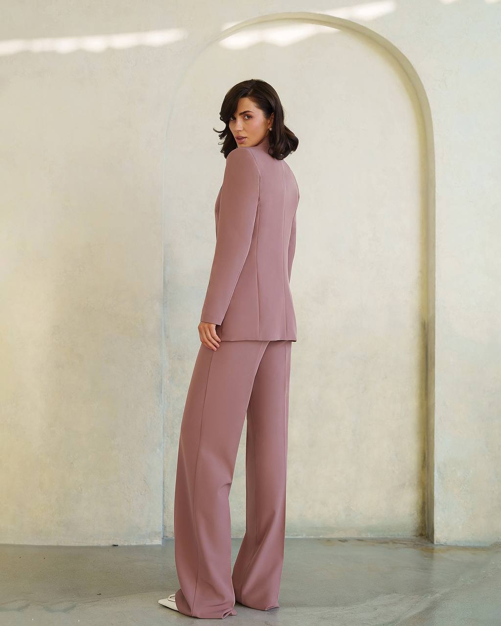 Woman wearing a mauve suit standing in a minimalistic room with a curved wall.