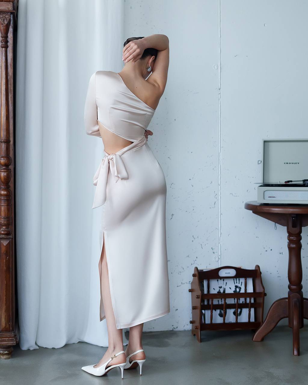 Woman in a beige dress standing in a room with a wooden side table and white curtain.