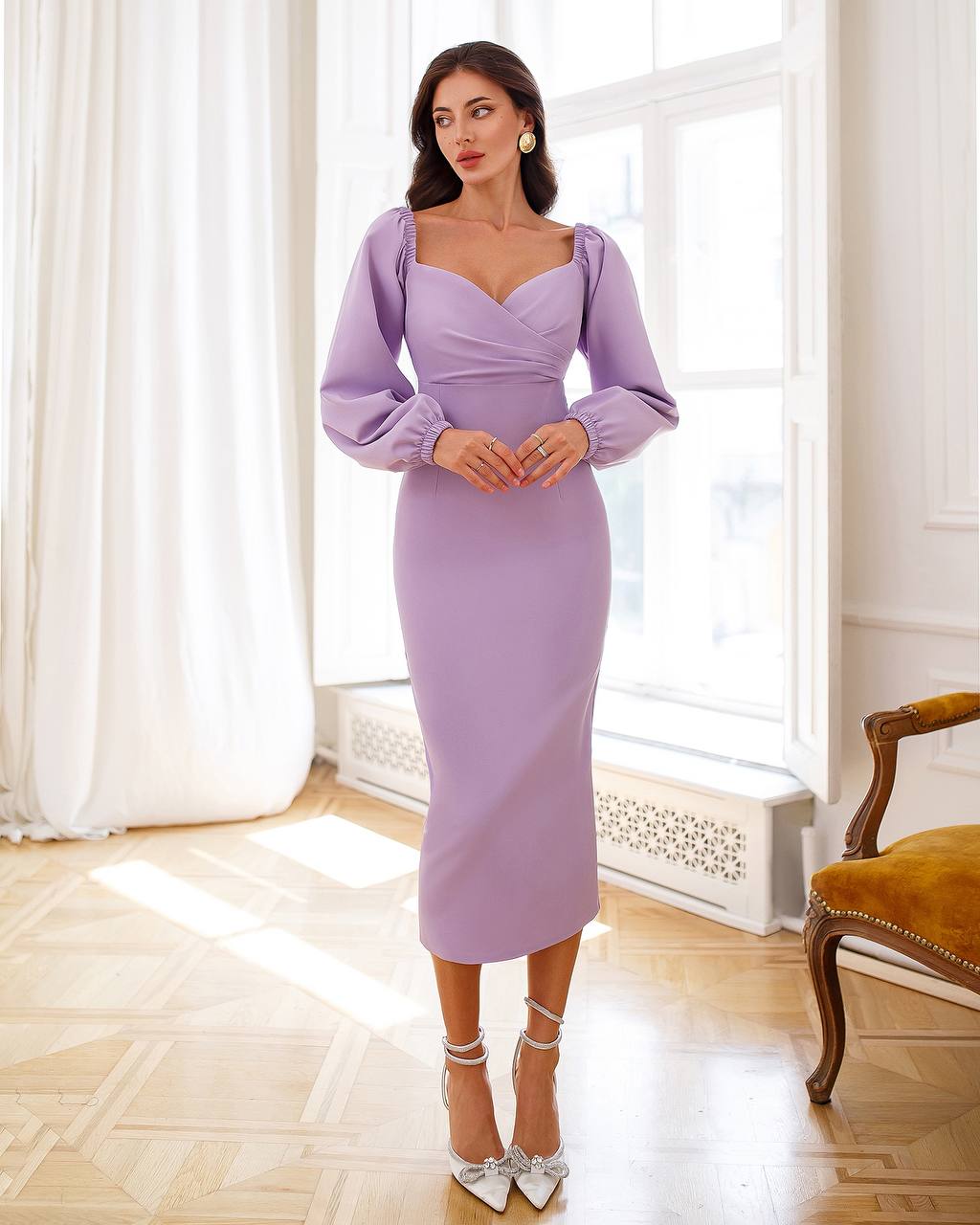 Woman wearing a lavender dress in a bright room with white curtains and a wooden floor.