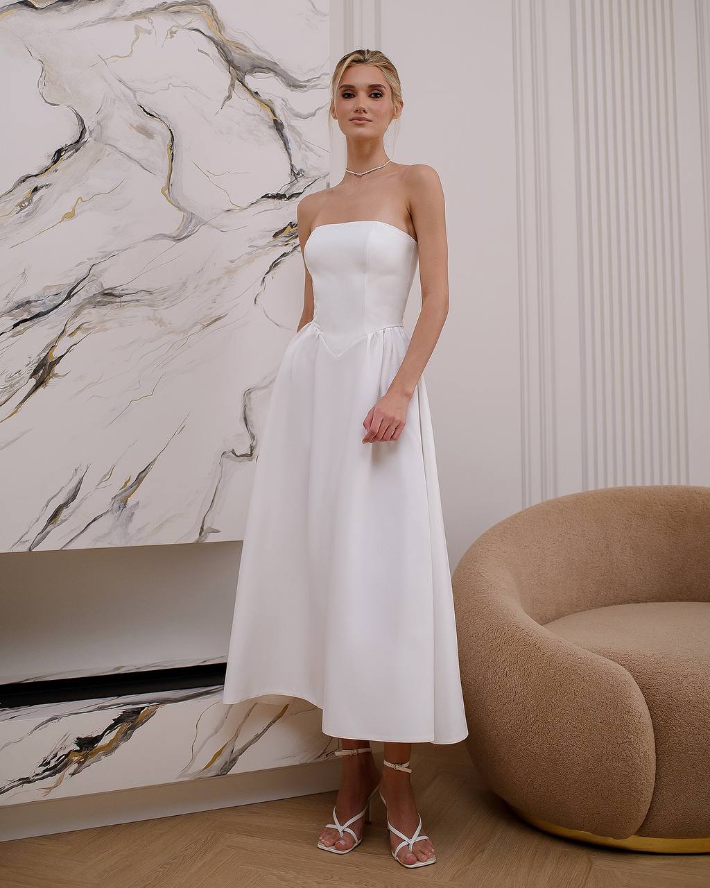Woman in a white strapless dress standing in a modern interior setting with a marble wall and brown chair.