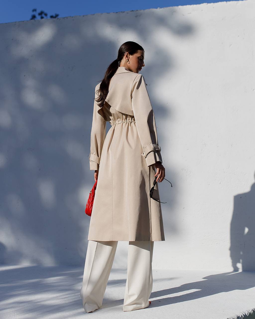 Woman in a beige trench coat standing against a white wall with blue sky.