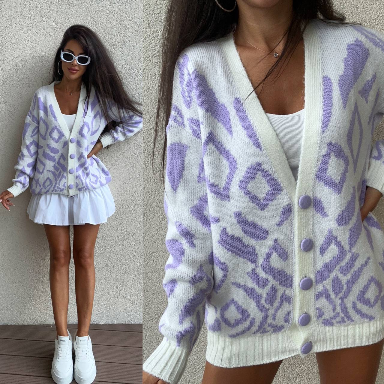 Woman wearing a white cardigan with purple geometric pattern, standing against a neutral wall.