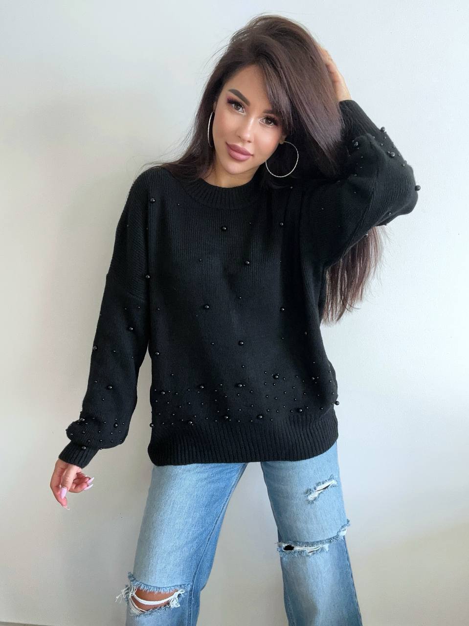 Woman wearing a black sweater with pearl embellishments and ripped jeans against a plain background