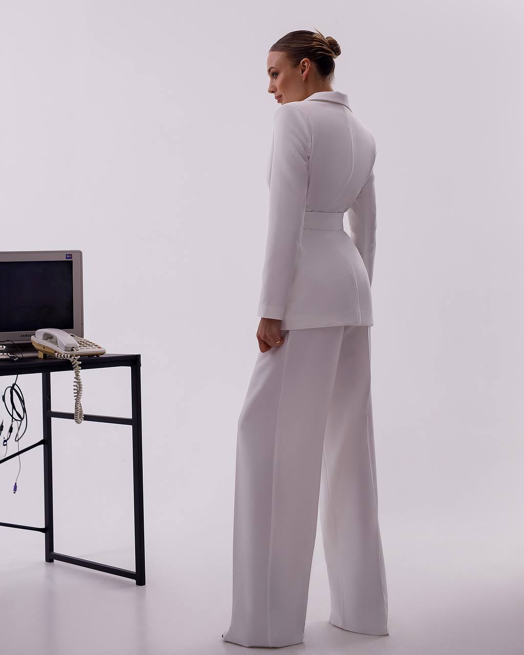 Woman in a white outfit standing in an office setting with a desk and computer.