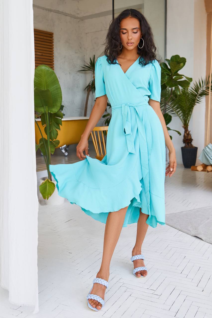 Woman in a light blue dress standing in a room with plants and furniture.