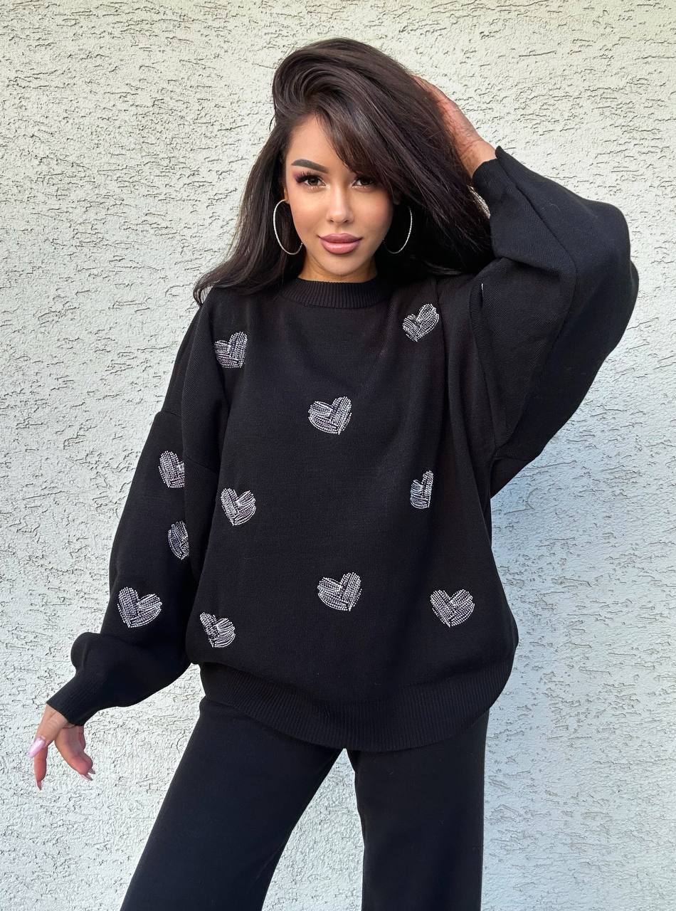 Woman wearing a black sweatshirt with heart patterns against a light gray wall.