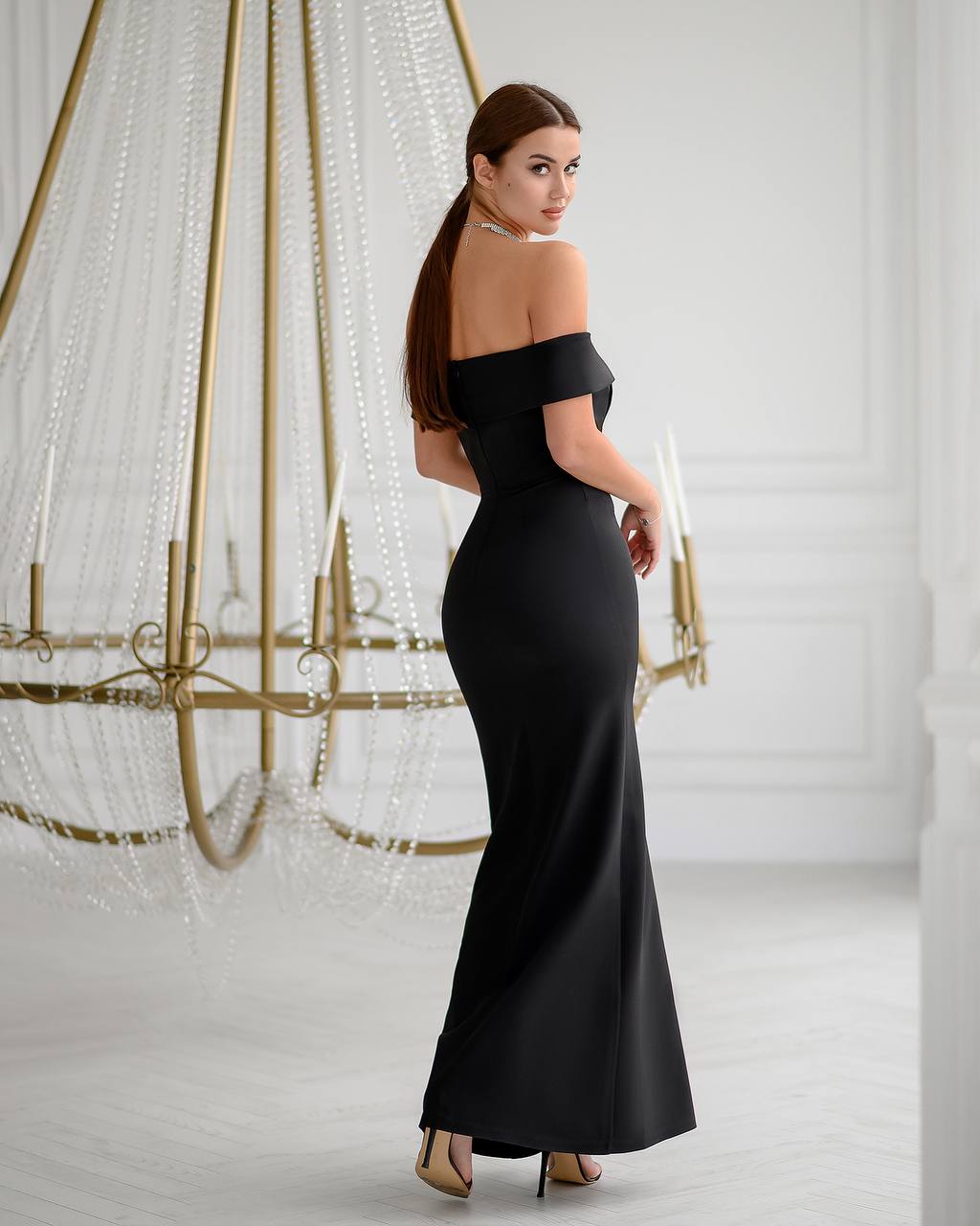 Woman in a black off-shoulder gown standing in front of a decorative chandelier.