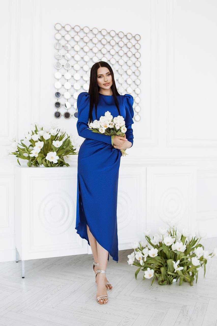 Woman in a blue dress holding flowers in a minimalistic setting with white walls and greenery.