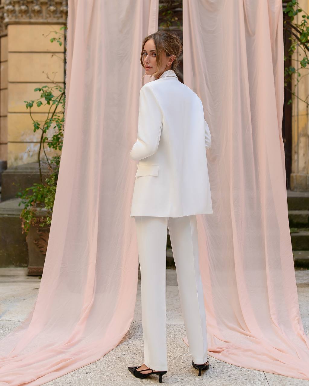 Woman in a white suit standing in front of pink curtains