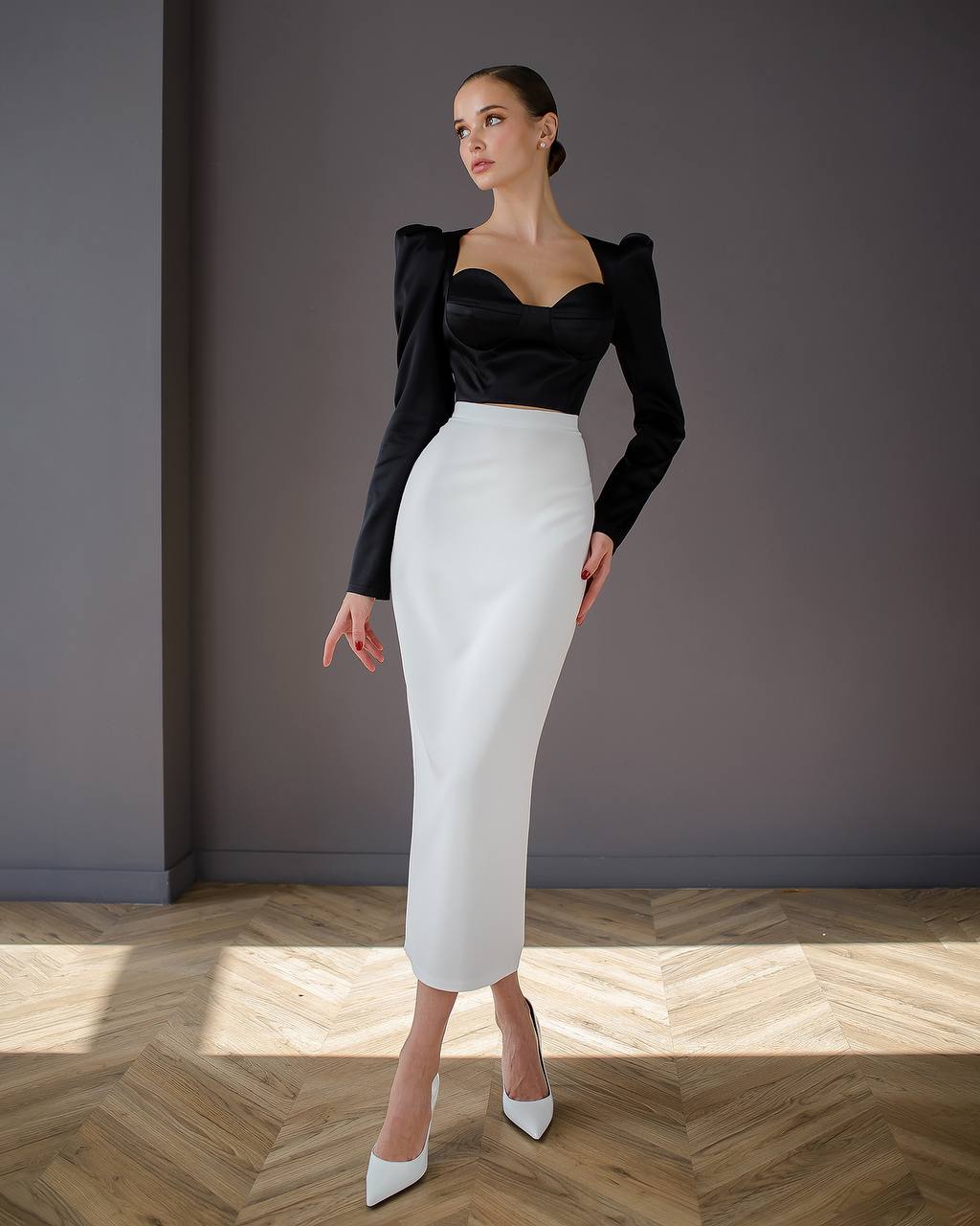 Woman wearing a black top and white skirt standing against a gray wall.