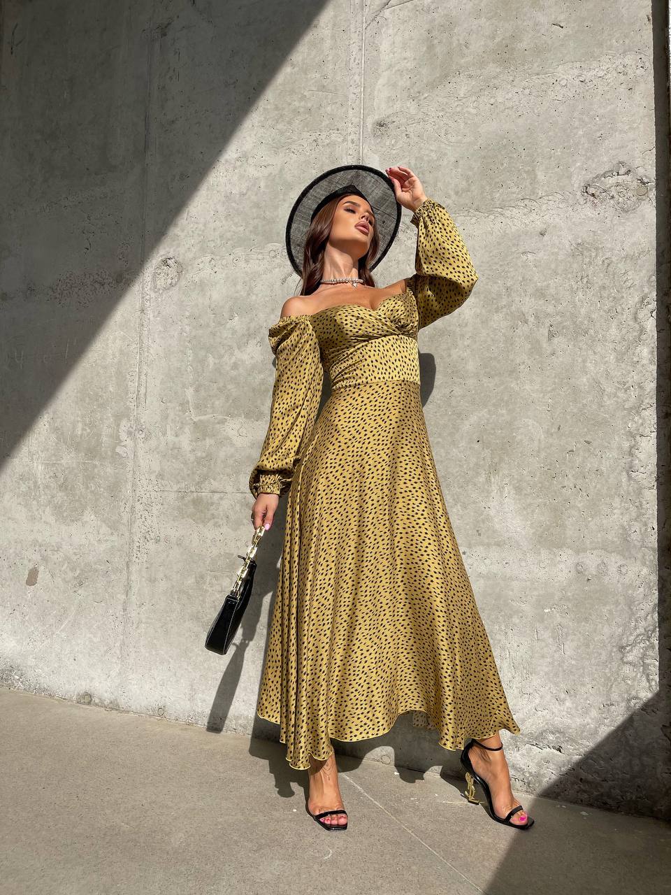 Woman in a yellow patterned dress and black hat against a concrete wall.