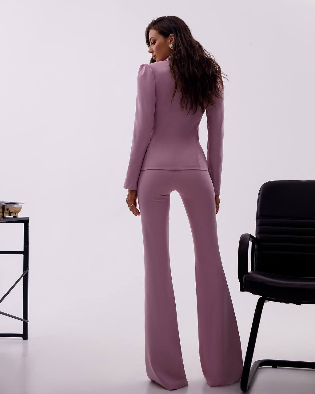 Woman wearing a mauve suit standing in a minimalistic room with a black chair and small table.