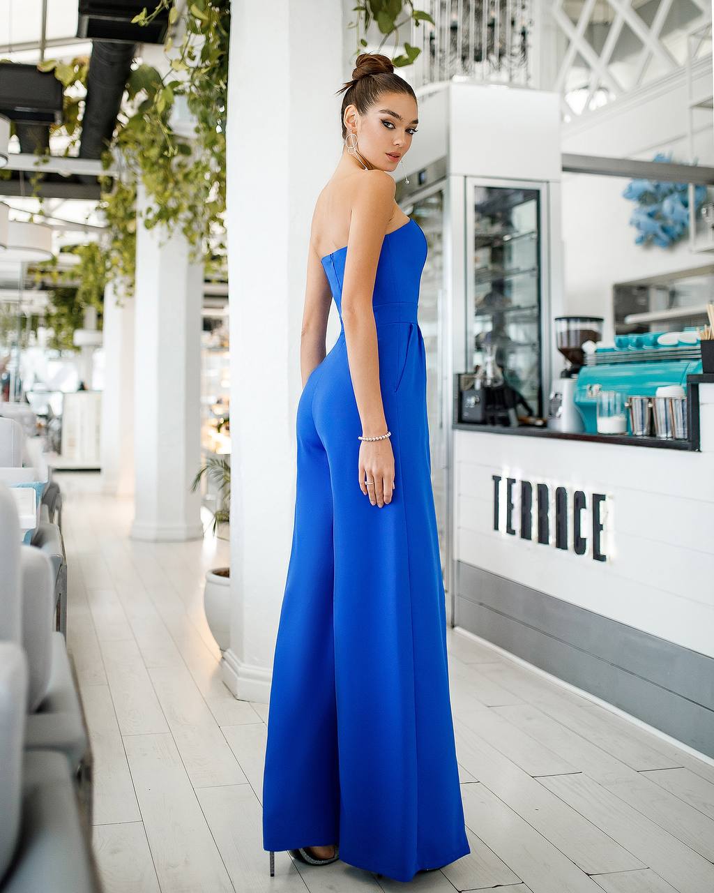 Woman in a blue strapless jumpsuit standing in a modern interior setting.